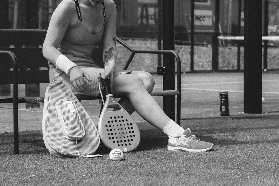 A padel player takes a break on a bench, resting her racket and ball on the ground beside her.