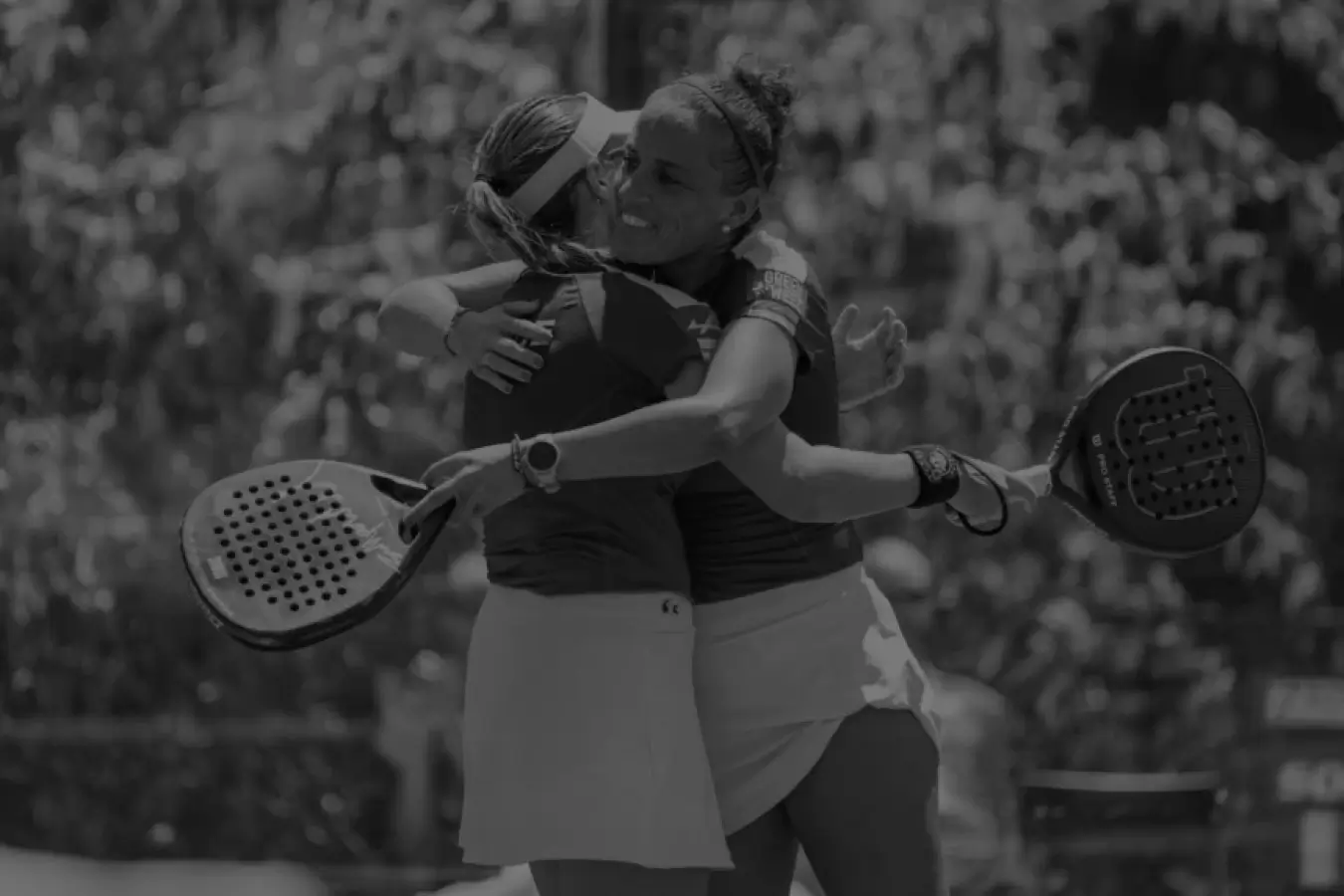 Two female padel players embrace each other in a celebratory hug after a match, both holding rackets and smiling warmly.