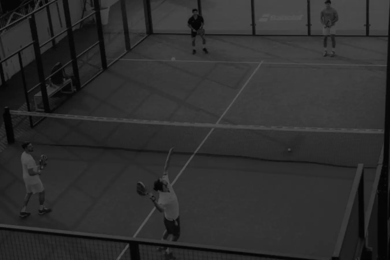 Four padel players on a court during a match, with one player preparing to serve.