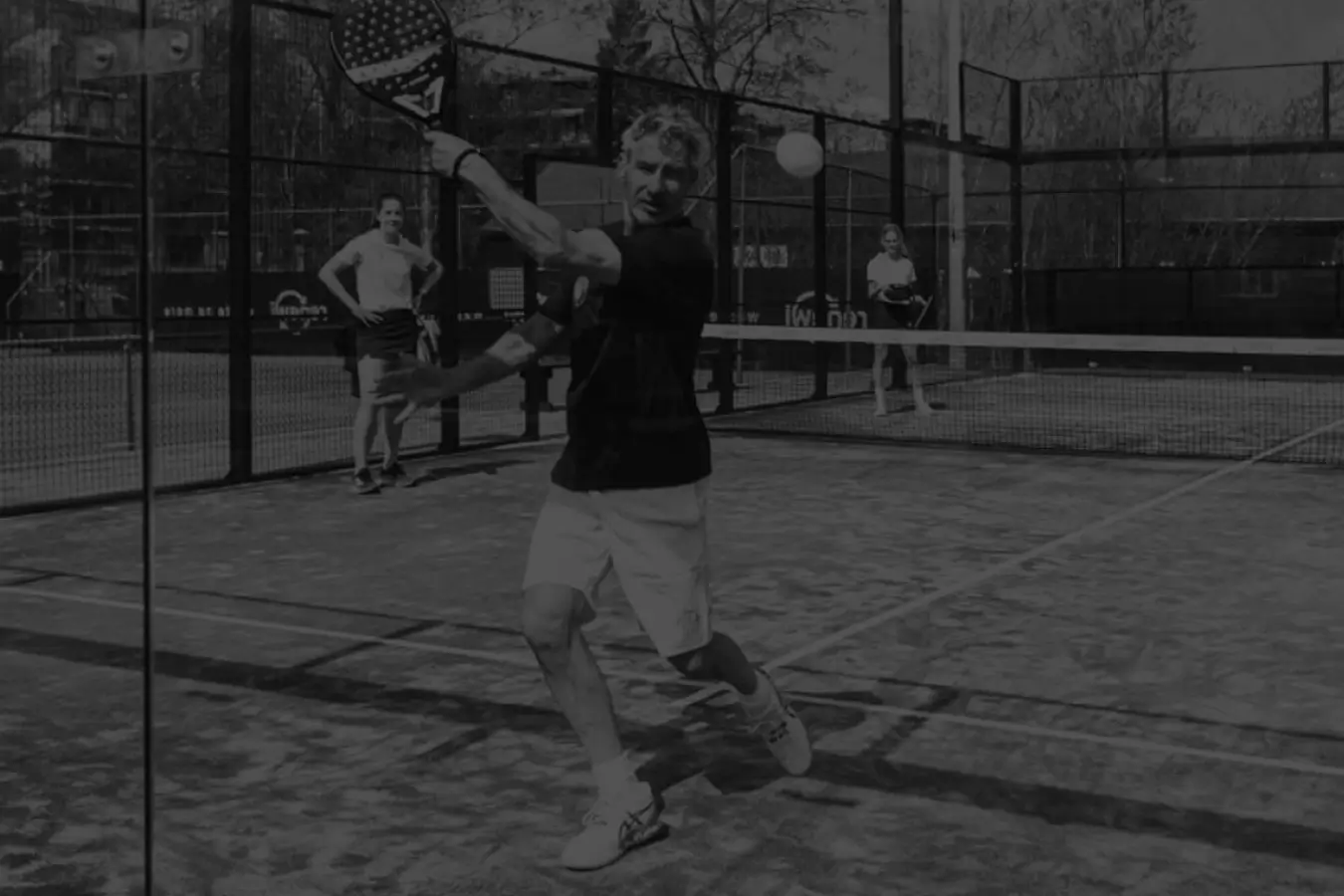 older man playing padel in black and white, hitting the ball on the glass