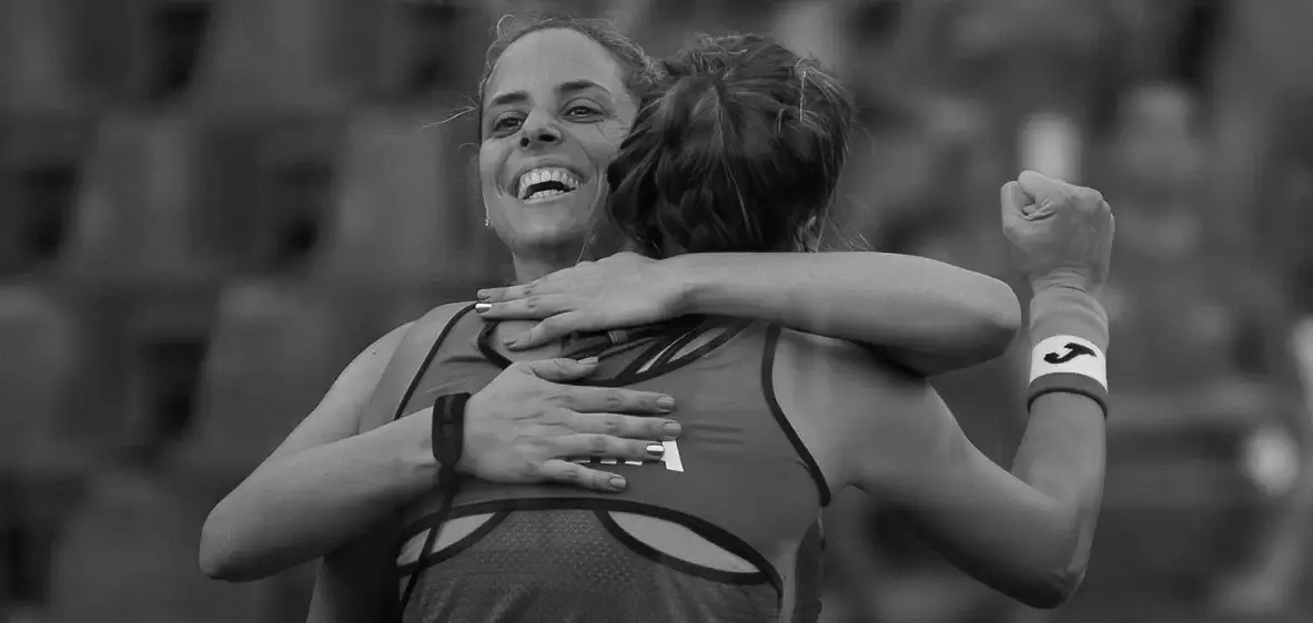 Two female padel players embracing in celebration after a match, one smiling with joy and the other raising a fist.