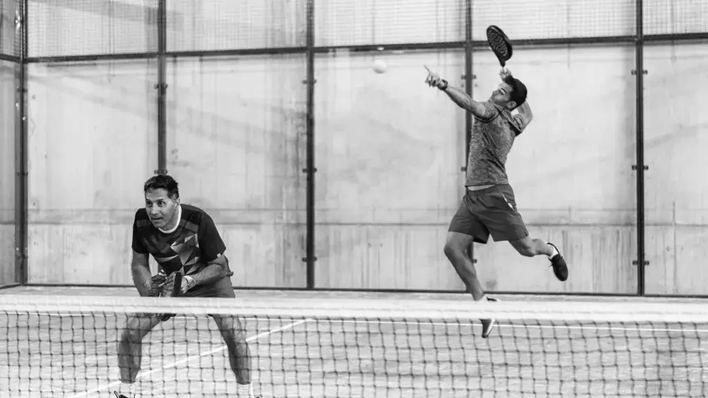 A player leaps into the air for a powerful smash while his partner waits at the net, capturing the fast-paced intensity of padel.